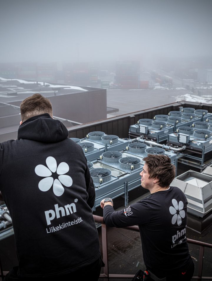 To vaktmestere fra PHM står på taket av en høy bygning og ser på utsikten over byen etter å ha utført vedlikehold av ventilasjonsenheter.