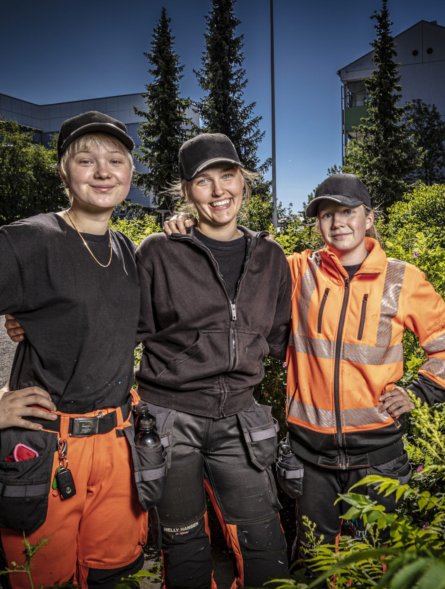 Tre smilende jenter poserer foran kamera etter å ha jobbet med vedlikehold av grøntarealene på en eiendom.